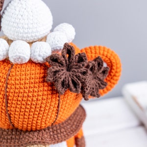 Puede incluir: Mu&ntilde;eca de gnomo de ganchillo hecha a mano con cuerpo naranja, bufanda marr&oacute;n y barba blanca. El gnomo tiene un sombrero blanco y est&aacute; adornado con adornos marrones en forma de estrella. El fondo es neutro.