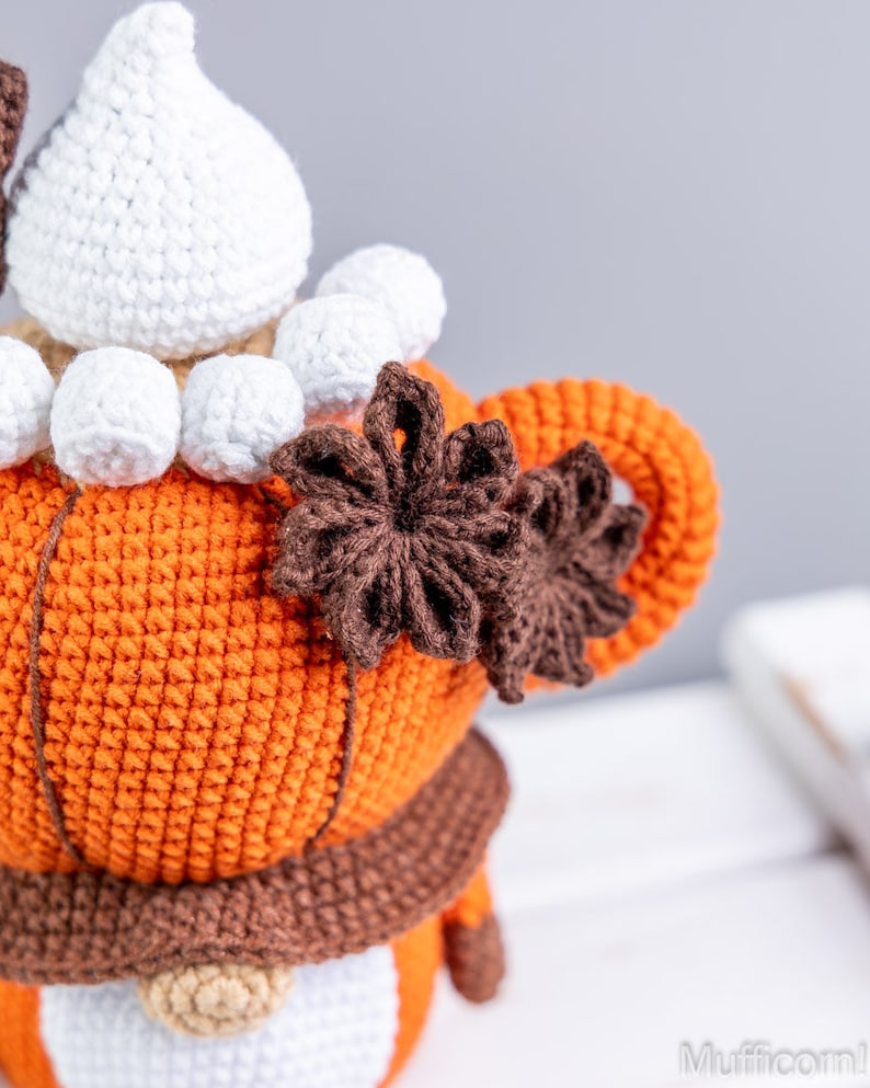 Puede incluir: Mu&ntilde;eca de gnomo de ganchillo hecha a mano con cuerpo naranja, bufanda marr&oacute;n y barba blanca. El gnomo tiene un sombrero blanco y est&aacute; adornado con adornos marrones en forma de estrella. El fondo es neutro.