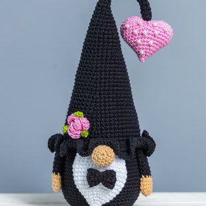 May include: A black crocheted gnome wearing a white tuxedo with a black bow tie and a pink heart on its head. The gnome has a pink flower on its hat and a brown beard.