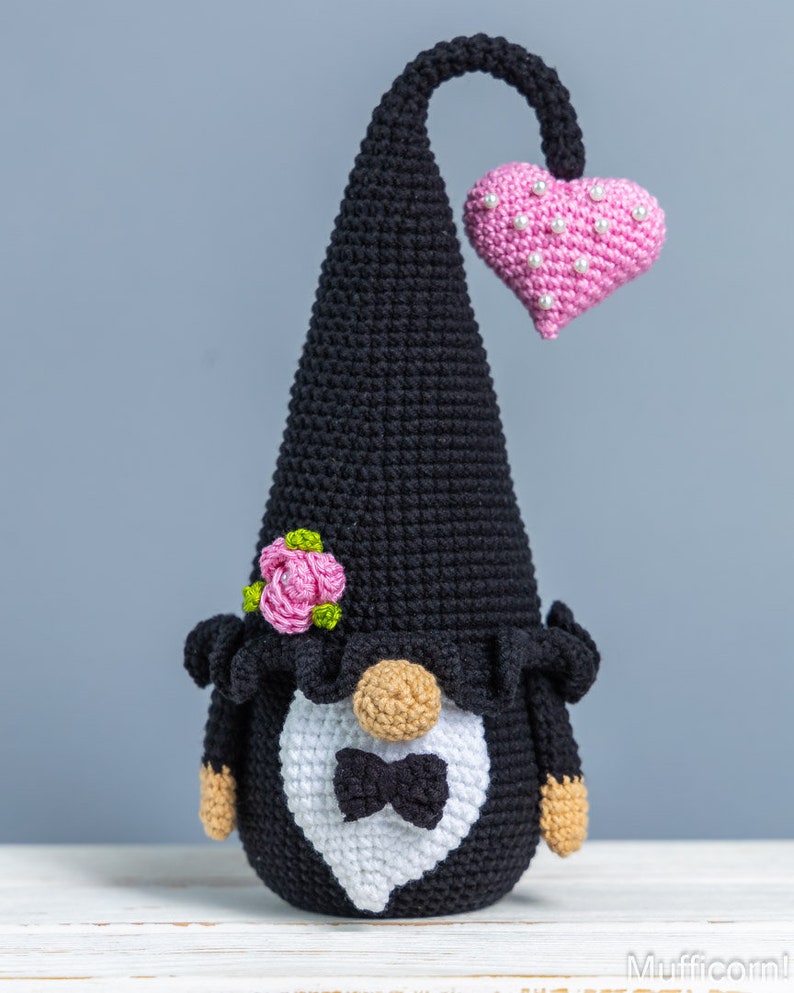 May include: A black crocheted gnome wearing a white tuxedo with a black bow tie and a pink heart on its head. The gnome has a pink flower on its hat and a brown beard.