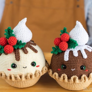 May include: Two crocheted Christmas pudding ornaments with white frosting, red holly berries, and green leaves. The ornaments have smiling faces with black eyes and brown cheeks.