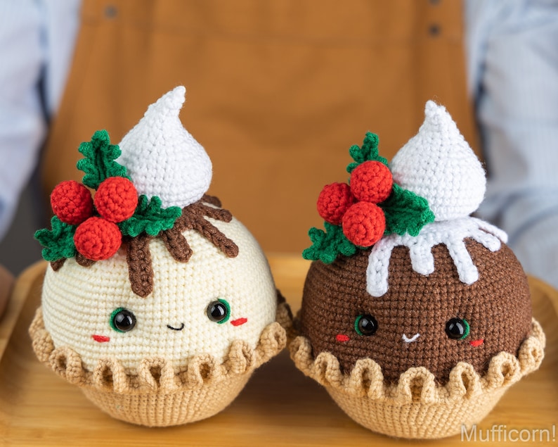 May include: Two crocheted Christmas pudding ornaments with white frosting, red holly berries, and green leaves. The ornaments have smiling faces with black eyes and brown cheeks.