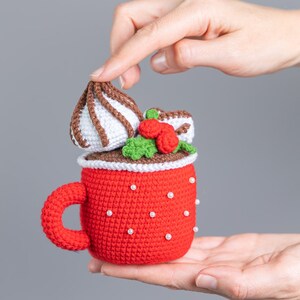 May include: A crocheted red mug with a white handle and white pearl accents. The mug is filled with a brown crocheted chocolate drink topped with a white crocheted marshmallow and green crocheted holly with red berries.