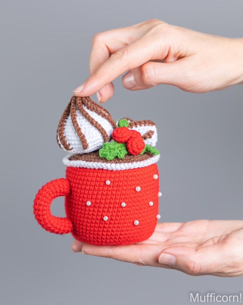 May include: A crocheted red mug with a white handle and white pearl accents. The mug is filled with a brown crocheted chocolate drink topped with a white crocheted marshmallow and green crocheted holly with red berries.