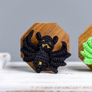 May include: A black crocheted bat with yellow eyes and a yellow star on its chest, displayed on a wooden octagon.