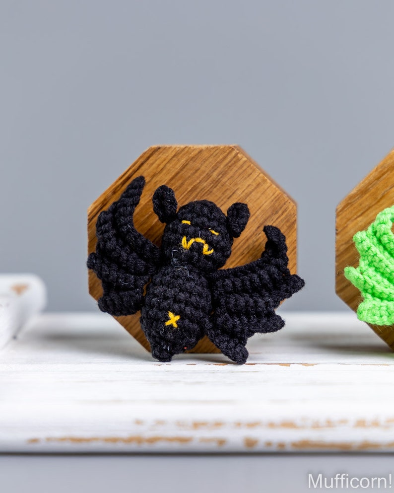 May include: A black crocheted bat with yellow eyes and a yellow star on its chest, displayed on a wooden octagon.