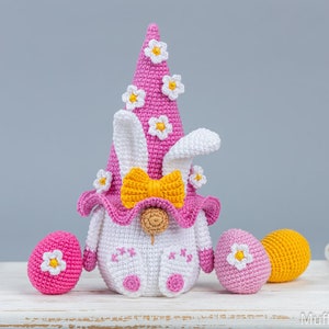 May include: A pink and white crocheted Easter bunny gnome with a yellow bow and white flowers on its hat. The gnome is sitting on a white surface with three crocheted Easter eggs in pink and yellow.