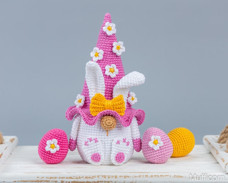 May include: A pink and white crocheted Easter bunny gnome with a yellow bow and white flowers on its hat. The gnome is sitting on a white surface with three crocheted Easter eggs in pink and yellow.
