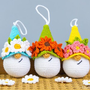 May include: Three crocheted gnomes with white faces and floral crowns. The gnome on the left has a blue hat with white daisies, the middle gnome has a green hat with orange flowers, and the gnome on the right has a yellow hat with pink flowers.