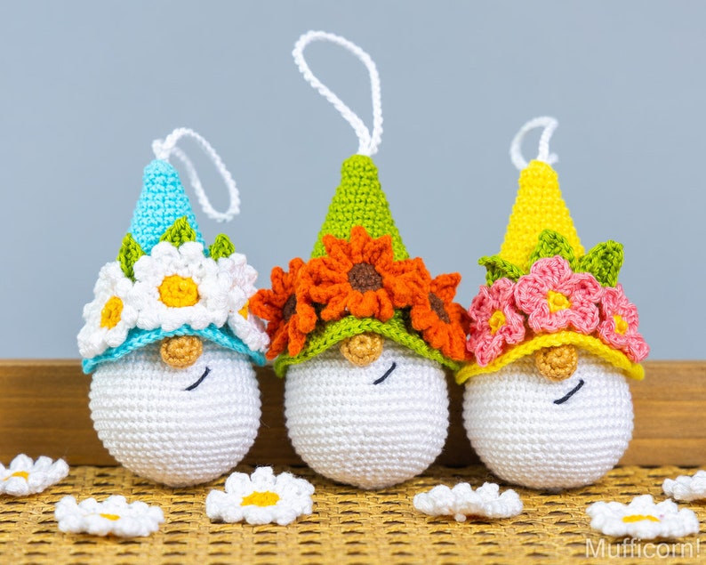 May include: Three crocheted gnomes with white faces and floral crowns. The gnome on the left has a blue hat with white daisies, the middle gnome has a green hat with orange flowers, and the gnome on the right has a yellow hat with pink flowers.