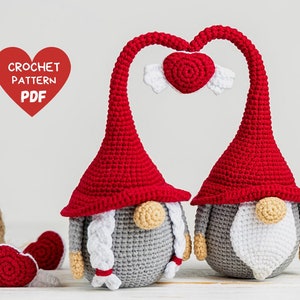 May include: Two crocheted gnomes with red hats and gray bodies, holding a red heart with white wings above their heads. The gnomes have white beards and noses. The image includes the text "CROCHET PATTERN PDF".