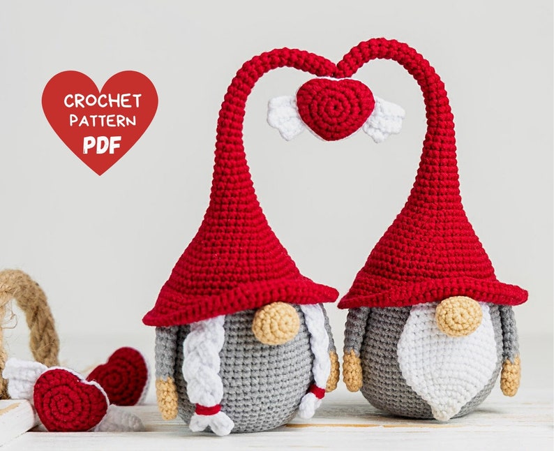 May include: Two crocheted gnomes with red hats and gray bodies, holding a red heart with white wings above their heads. The gnomes have white beards and noses. The image includes the text "CROCHET PATTERN PDF".