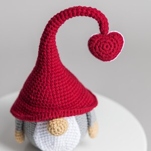 May include: A crocheted gnome with a red hat and a heart-shaped top. The gnome has a white beard and gray body.