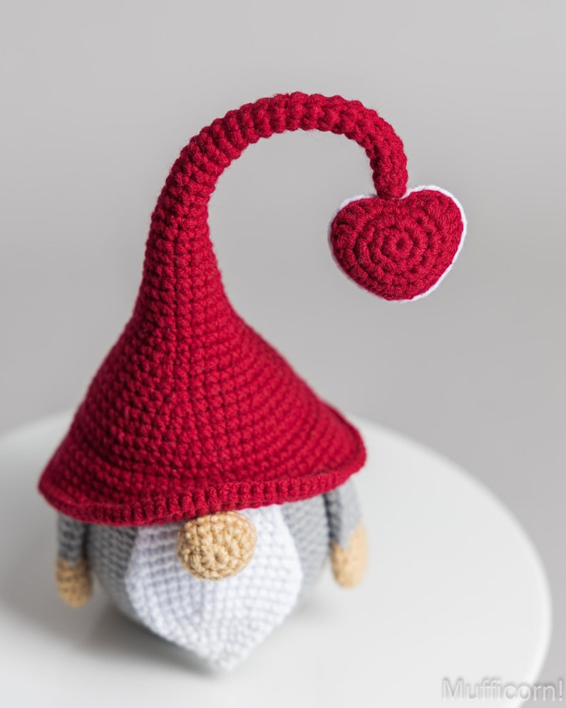 May include: A crocheted gnome with a red hat and a heart-shaped top. The gnome has a white beard and gray body.