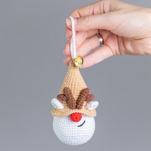 May include: A crocheted reindeer ornament with a brown hat, red nose, and brown antlers. The ornament is white with a brown and white striped hat. The ornament has a white loop for hanging.