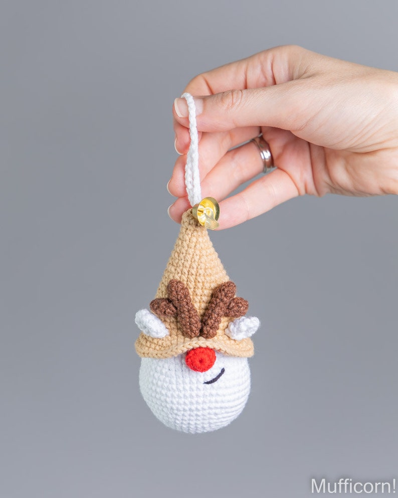 May include: A crocheted reindeer ornament with a brown hat, red nose, and brown antlers. The ornament is white with a brown and white striped hat. The ornament has a white loop for hanging.