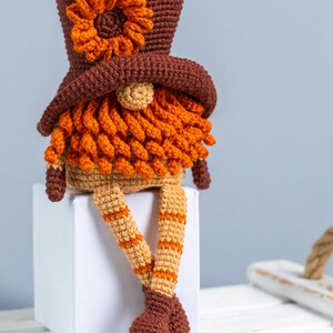 May include: A crocheted gnome with a brown hat, orange beard, and tan legs. The gnome is sitting on a white cube.