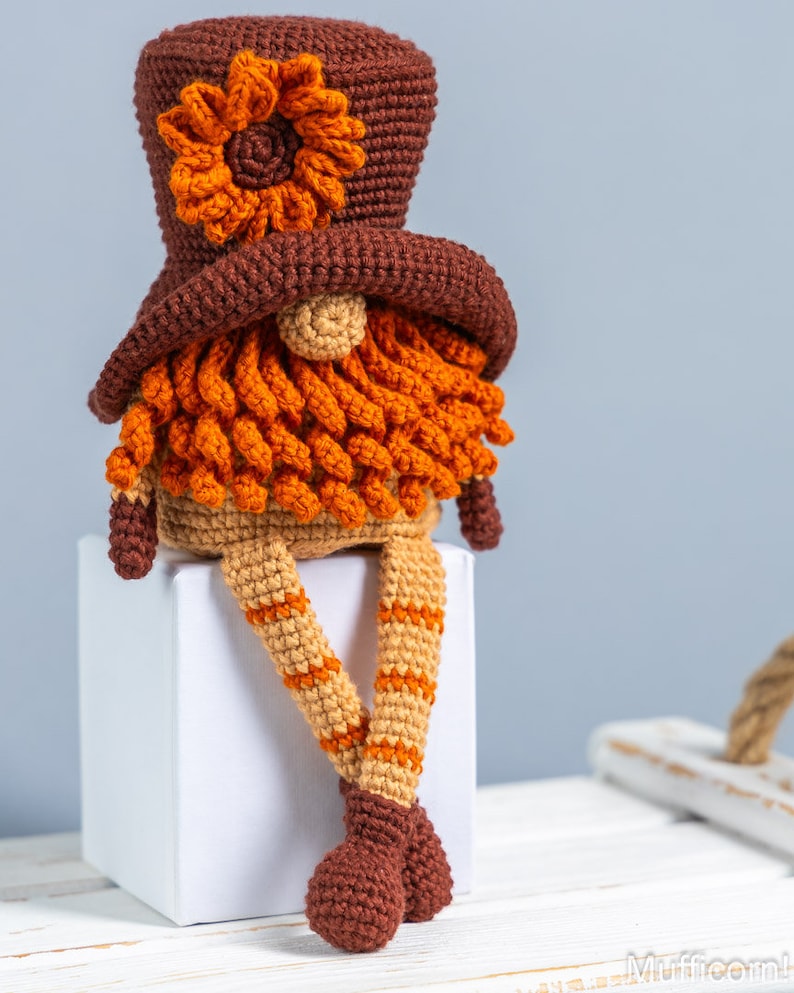 May include: A crocheted gnome with a brown hat, orange beard, and tan legs. The gnome is sitting on a white cube.