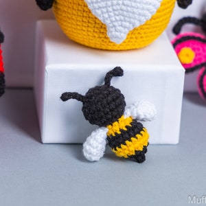 May include: A crocheted bee toy with black and yellow stripes, white wings, and a black head. The bee is sitting on a white surface.