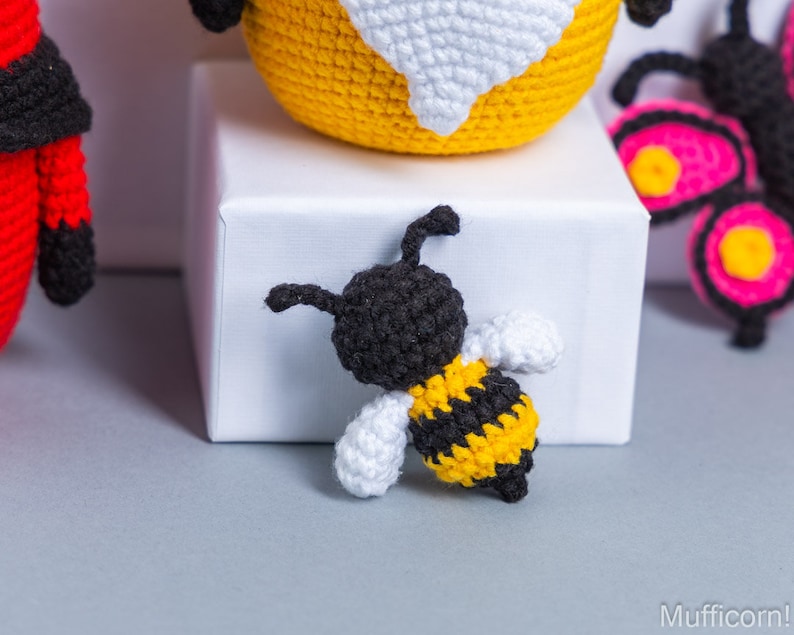 May include: A crocheted bee toy with black and yellow stripes, white wings, and a black head. The bee is sitting on a white surface.