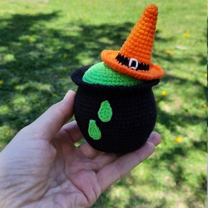 May include: A crocheted witch's cauldron with a green cauldron and a black rim. The cauldron has a green liquid inside and is topped with an orange witch's hat with a black buckle.