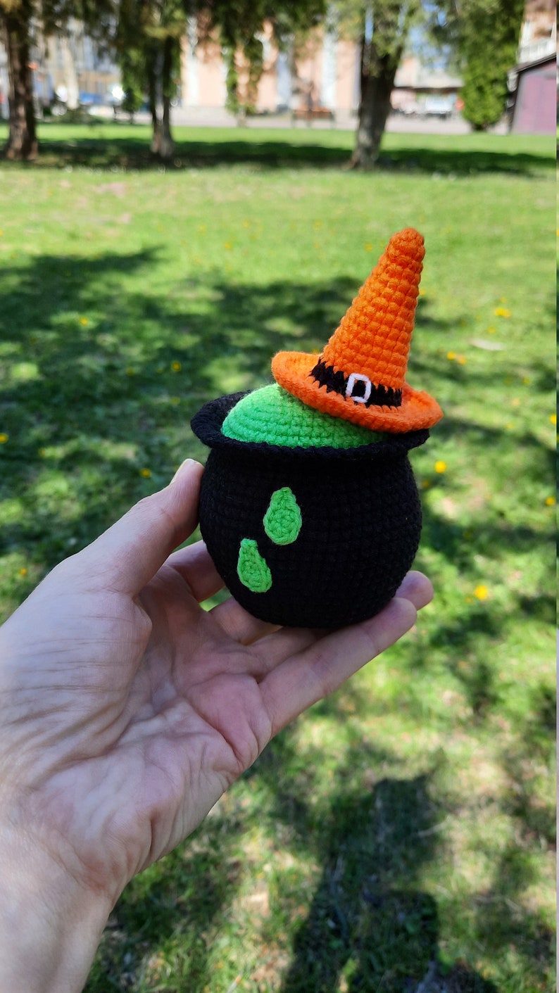 May include: A crocheted witch's cauldron with a green cauldron and a black rim. The cauldron has a green liquid inside and is topped with an orange witch's hat with a black buckle.