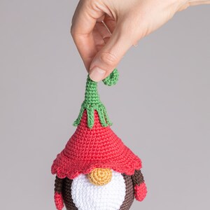 May include: A crocheted gnome ornament with a red hat, white beard, and brown body. The gnome has a green pointed hat with a curled tip and wears brown mittens.