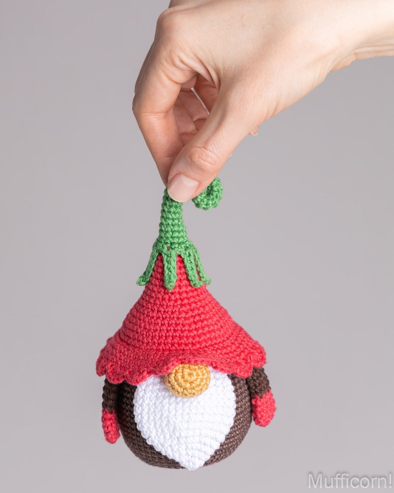 May include: A crocheted gnome ornament with a red hat, white beard, and brown body. The gnome has a green pointed hat with a curled tip and wears brown mittens.