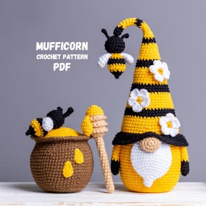 May include: A crocheted bee gnome with a yellow and black striped hat, white flowers, and a white beard. The gnome is holding a honey dipper and a pot of honey with a bee on top. The text "MUFFICORN CROCHET PATTERN PDF" is in the top left corner of the image.