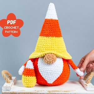 May include: Crochet pattern for a candy corn gnome. The gnome is wearing a white, orange, and yellow hat and has a white beard. The gnome is holding a small candy corn in its hand. The pattern is available as a PDF download.