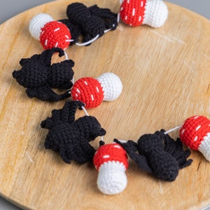 May include: A garland of crocheted black bats and red and white mushrooms. The garland is strung together with white yarn and is displayed on a wooden surface.