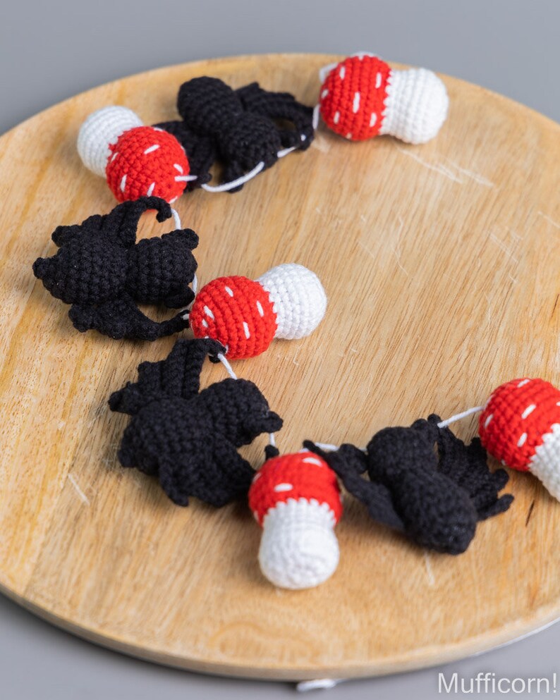 May include: A garland of crocheted black bats and red and white mushrooms. The garland is strung together with white yarn and is displayed on a wooden surface.