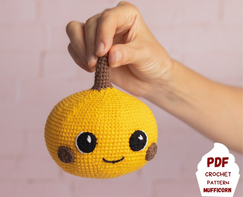 May include: A crocheted yellow pumpkin with a brown stem and a smiling face. The pumpkin has black eyes and a brown nose. The crocheted pumpkin is being held by a hand.