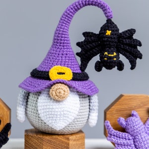 Puede incluir: Un gnomo de crochet morado que lleva un sombrero de bruja con un cintur&oacute;n negro y una hebilla amarilla. El gnomo tiene una barba blanca y sostiene un murci&eacute;lago de crochet negro. El gnomo est&aacute; sentado en un soporte de madera.