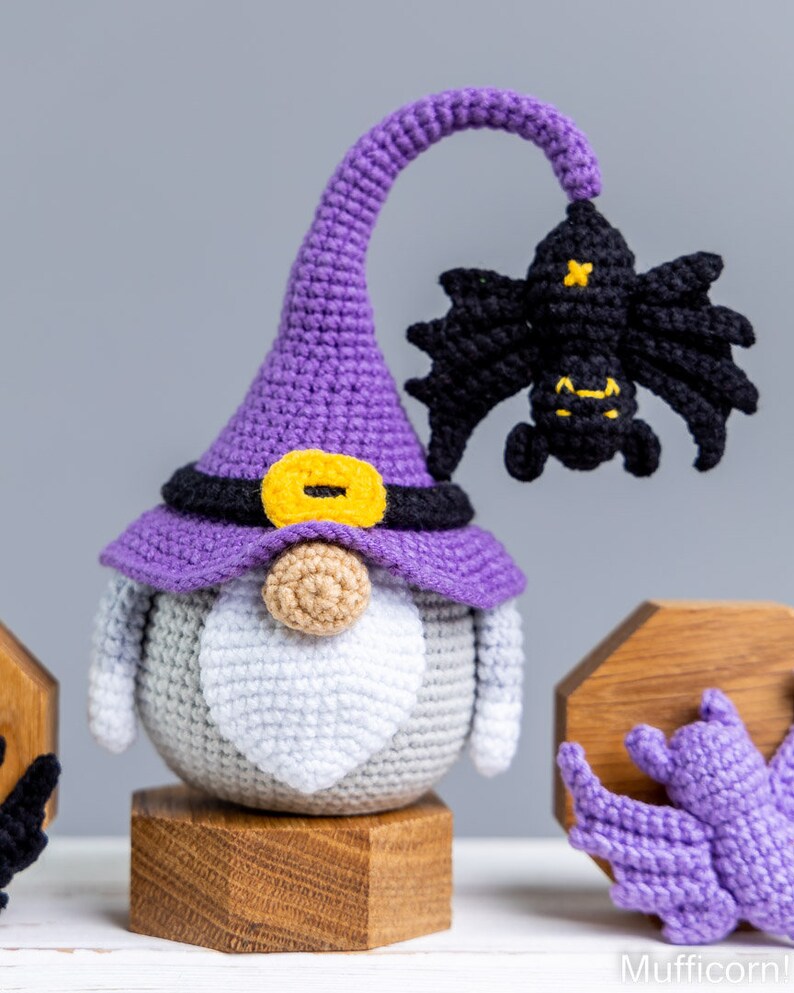 Puede incluir: Un gnomo de crochet morado que lleva un sombrero de bruja con un cintur&oacute;n negro y una hebilla amarilla. El gnomo tiene una barba blanca y sostiene un murci&eacute;lago de crochet negro. El gnomo est&aacute; sentado en un soporte de madera.