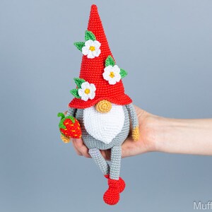 Op de afbeelding: Een gehaakte gnome met een rode muts, witte bloemen en een groen bladontwerp. De gnome houdt een rode aardbei vast en heeft grijze benen en rode schoenen.