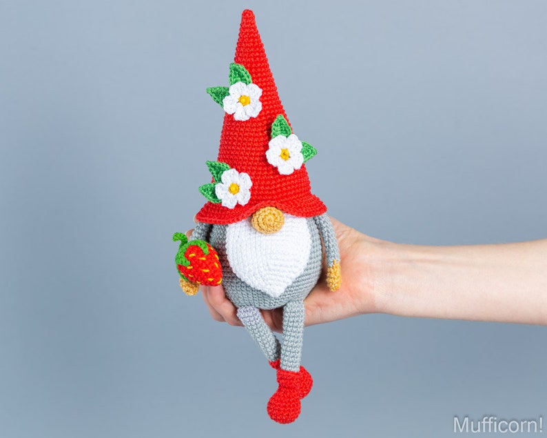 Op de afbeelding: Een gehaakte gnome met een rode muts, witte bloemen en een groen bladontwerp. De gnome houdt een rode aardbei vast en heeft grijze benen en rode schoenen.