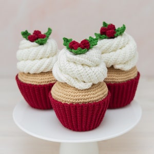 May include: Three crocheted cupcakes decorated with white frosting and red holly berries. The cupcakes are displayed on a white cake stand.