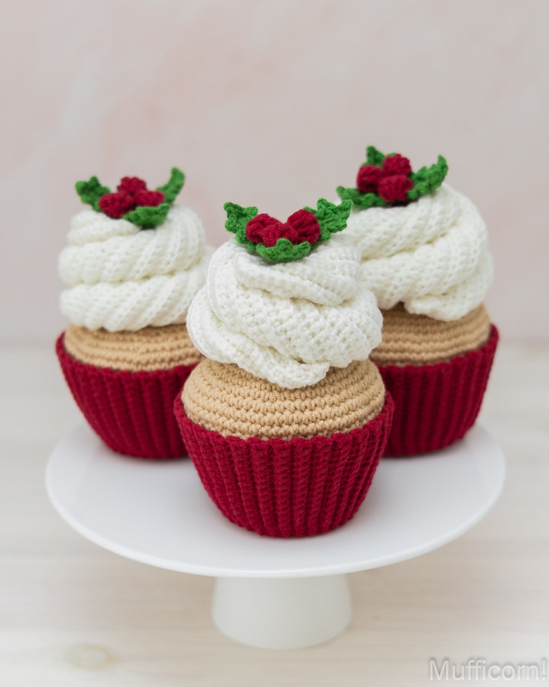 May include: Three crocheted cupcakes decorated with white frosting and red holly berries. The cupcakes are displayed on a white cake stand.