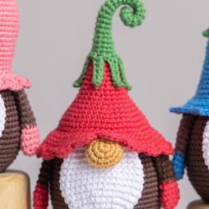 May include: Three crocheted gnomes with different coloured hats. The gnome in the centre has a red hat with a green curled top, a white beard, and a brown body. The gnome on the left has a pink hat and the gnome on the right has a blue hat.
