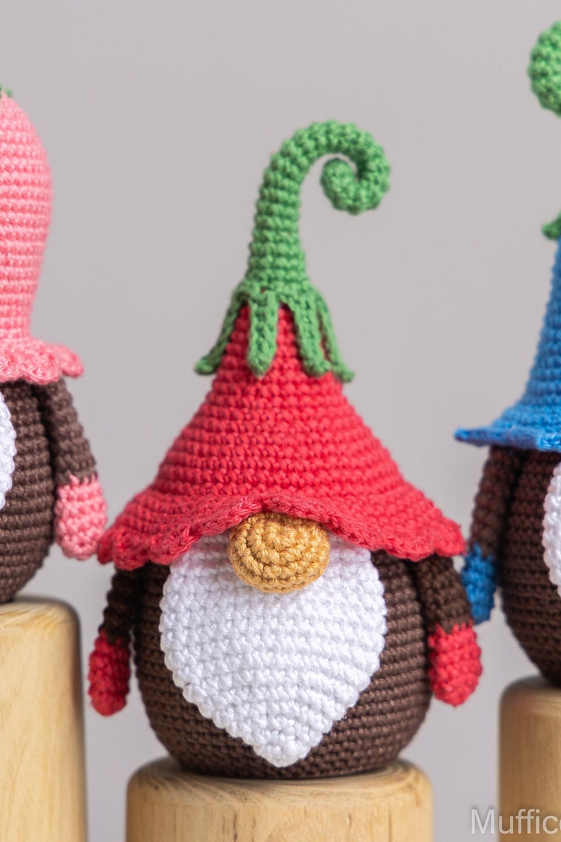 May include: Three crocheted gnomes with different coloured hats. The gnome in the centre has a red hat with a green curled top, a white beard, and a brown body. The gnome on the left has a pink hat and the gnome on the right has a blue hat.