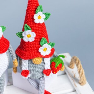 Op de afbeelding: Twee gehaakte gnomen met rode mutsen en witte bloemen. De gnome rechts houdt een rode gehaakte aardbei vast.