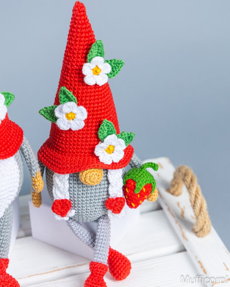 Op de afbeelding: Twee gehaakte gnomen met rode mutsen en witte bloemen. De gnome rechts houdt een rode gehaakte aardbei vast.
