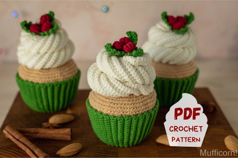 May include: Three crocheted Christmas cupcake ornaments with green icing and red holly berries. The ornaments are sitting on a wooden surface with cinnamon sticks and almonds. A white sign with the text "PDF CROCHET PATTERN" is in the foreground.