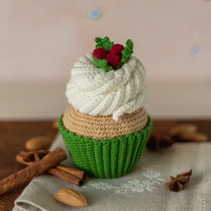 May include: A crocheted cupcake with white frosting and green icing, topped with red berries and green leaves. The cupcake is sitting on a wooden surface with cinnamon sticks and star anise.