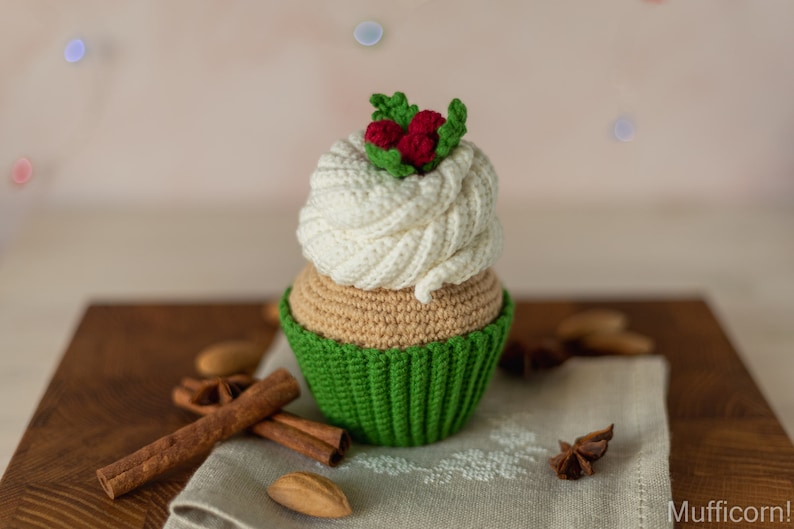 May include: A crocheted cupcake with white frosting and green icing, topped with red berries and green leaves. The cupcake is sitting on a wooden surface with cinnamon sticks and star anise.