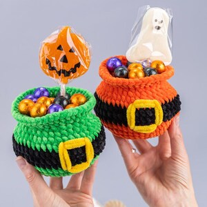 May include: Two crocheted pots filled with wrapped candies. One pot is green with a black belt and yellow buckle, holding a pumpkin lollipop. The other is orange with a black belt and yellow buckle, holding a ghost-shaped treat in clear plastic.