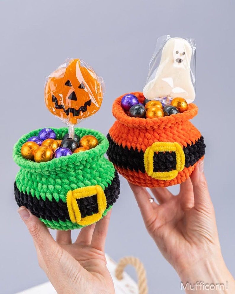 May include: Two crocheted pots filled with wrapped candies. One pot is green with a black belt and yellow buckle, holding a pumpkin lollipop. The other is orange with a black belt and yellow buckle, holding a ghost-shaped treat in clear plastic.