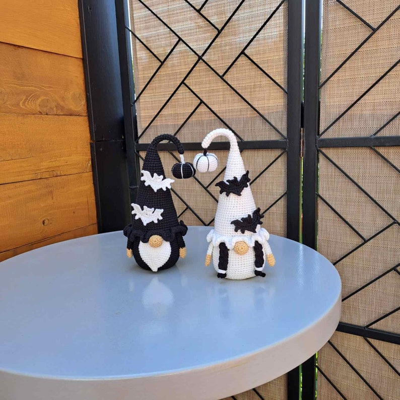 May include: Two crocheted gnomes with black and white accents. One gnome is wearing a black hat with a white bat and the other is wearing a white hat with a black bat. Both gnomes are holding a small crocheted pumpkin.