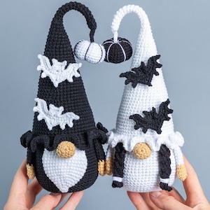 May include: Two crocheted gnomes dressed in black and white with bat and pumpkin accents. The gnomes are holding a small crocheted pumpkin.